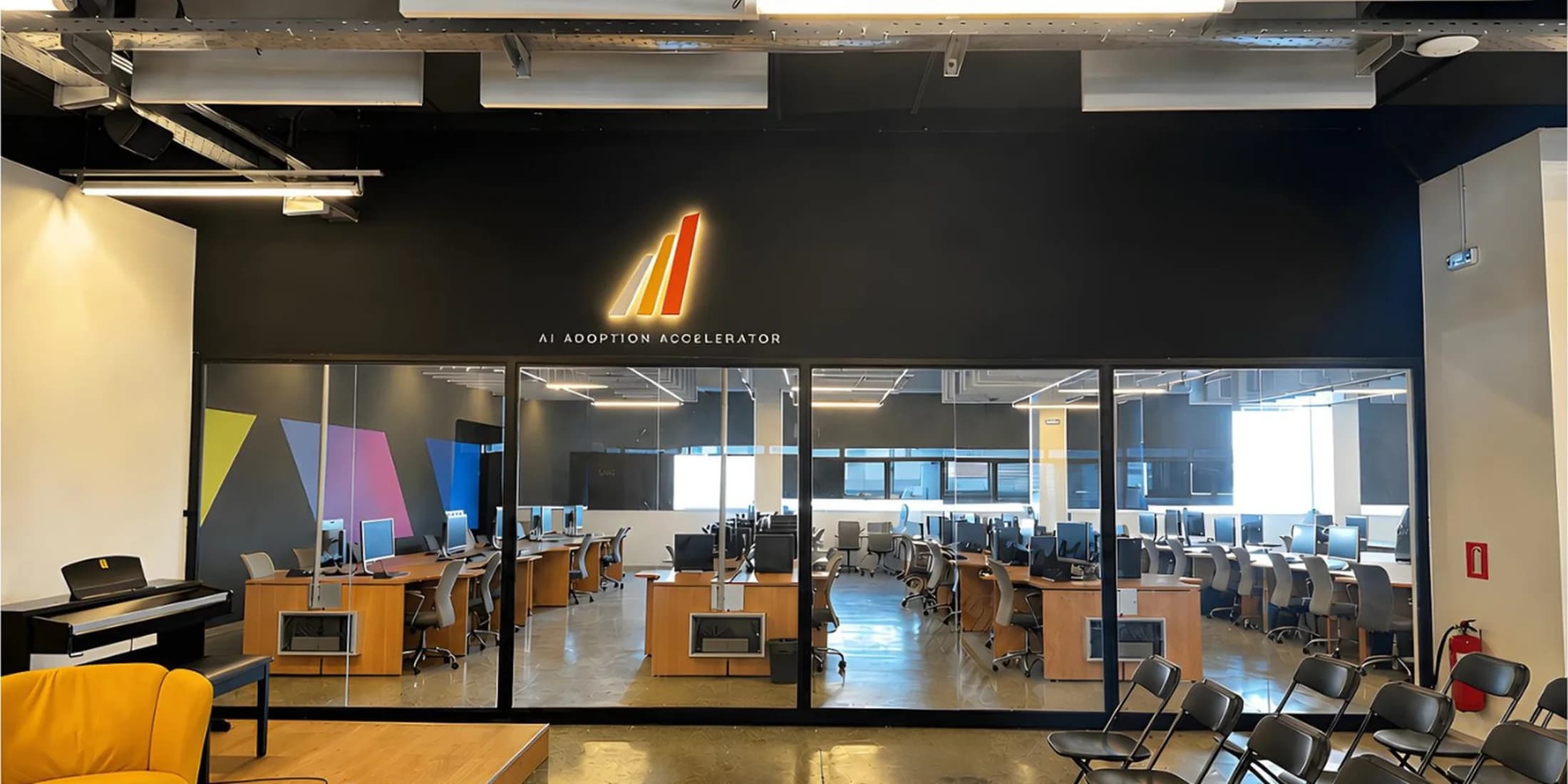 An open-plan workspace seen through a glazed partition, rows of desks and monitors below an illuminated A³ sign reading AI Adoption Accelerator.